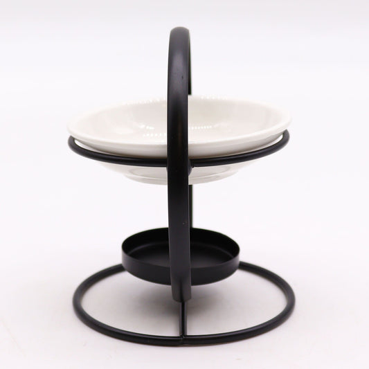 A modern essential oil burner featuring a circular black metal frame holding a shallow white ceramic bowl, with a small tealight holder at the base.