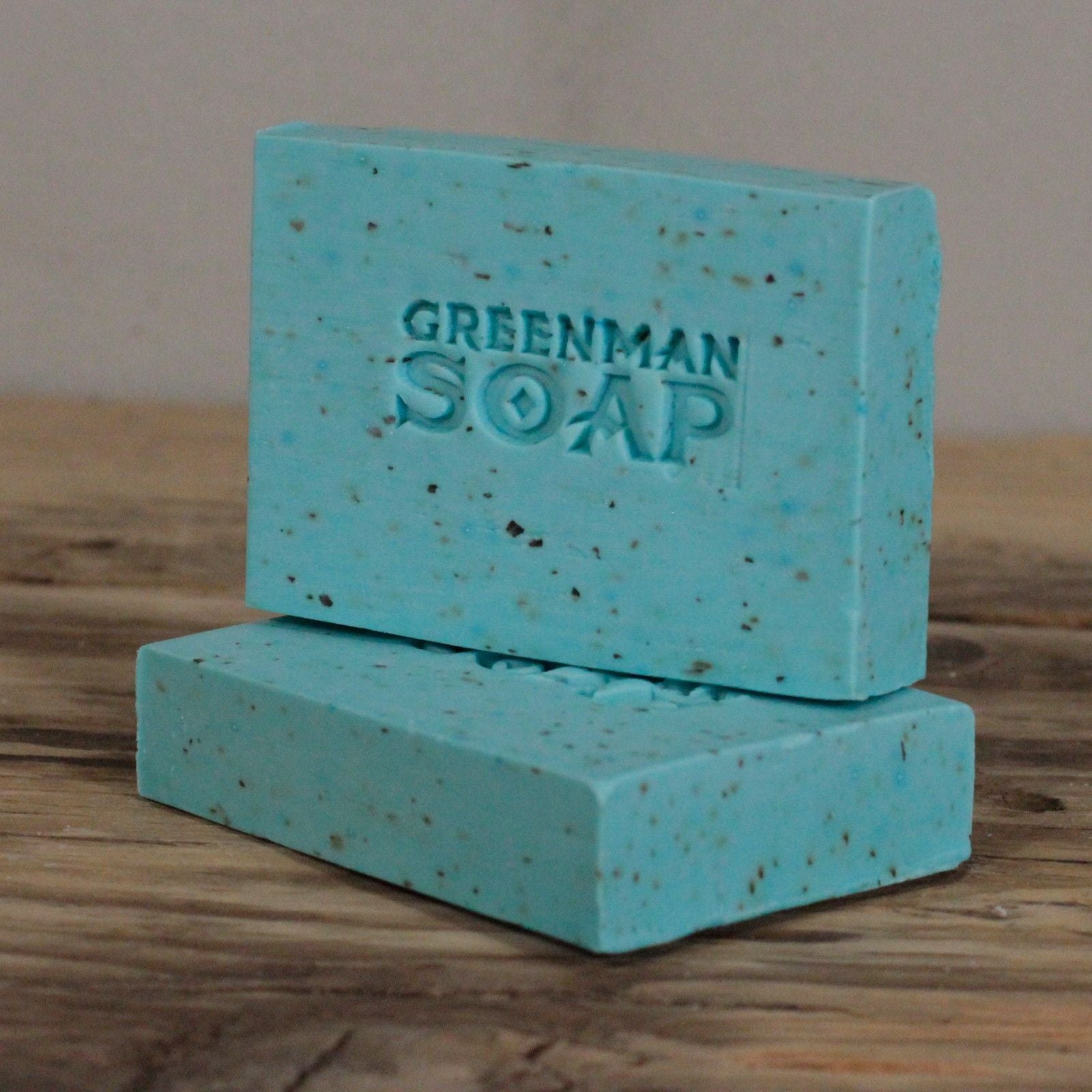 Handcrafted light blue Greenman artisan soap slices featuring dried mint leaves, presented on a natural wood surface.