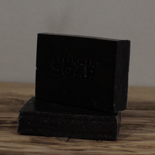 Handcrafted black charcoal Greenman artisan soap slices, presented on a natural wood surface.